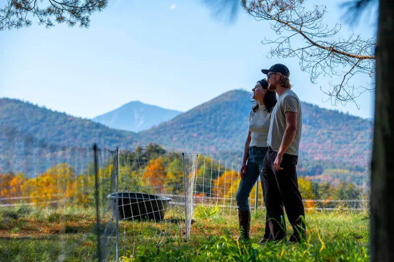 A couple standing in front of a farm with fall views and mountains in the background.