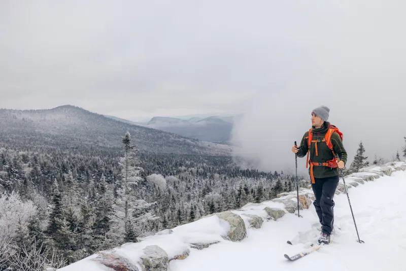 A man cross-country skis up a mountain trail with a view of trees down below.