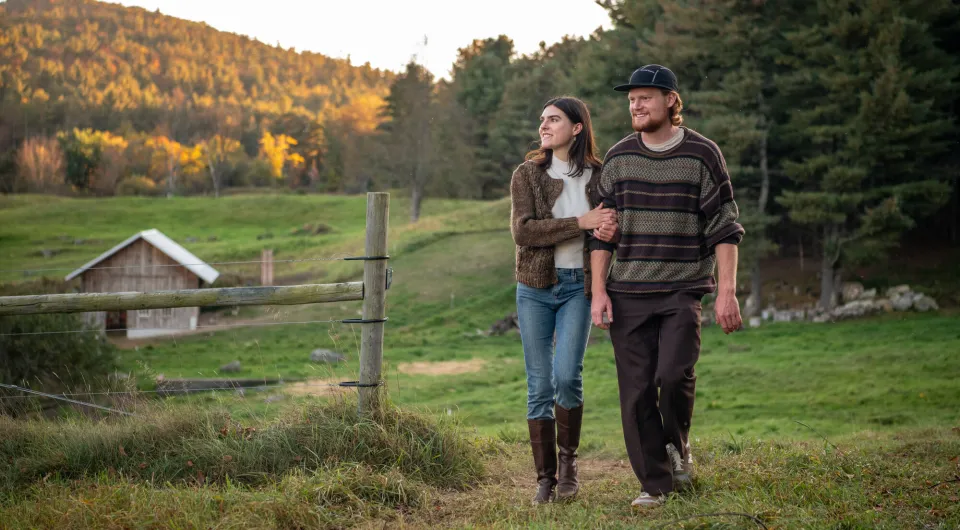 A couple walks arm in arm through a farm field with fall hues in the mountains