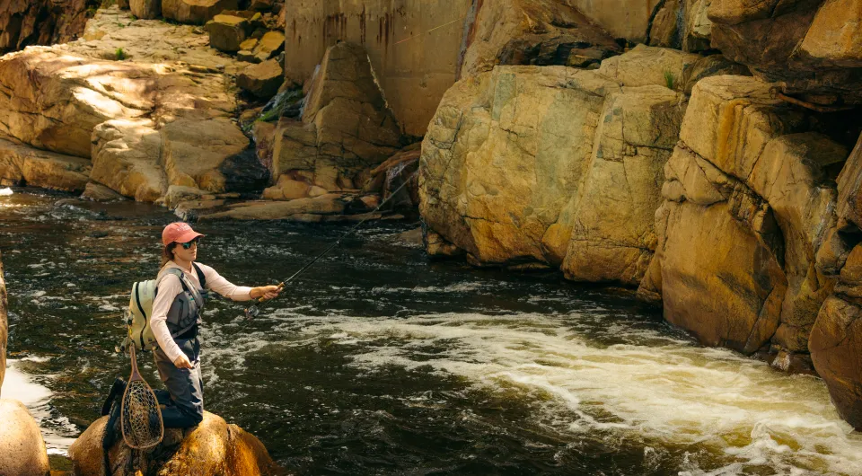 A woman in waders and pink hat fly fishes craggy section of Ausable River