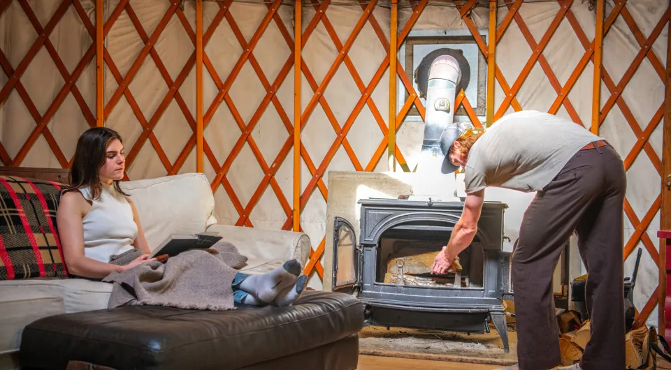 A man loads up a fireplace in a yurt while a woman reads on the couch.