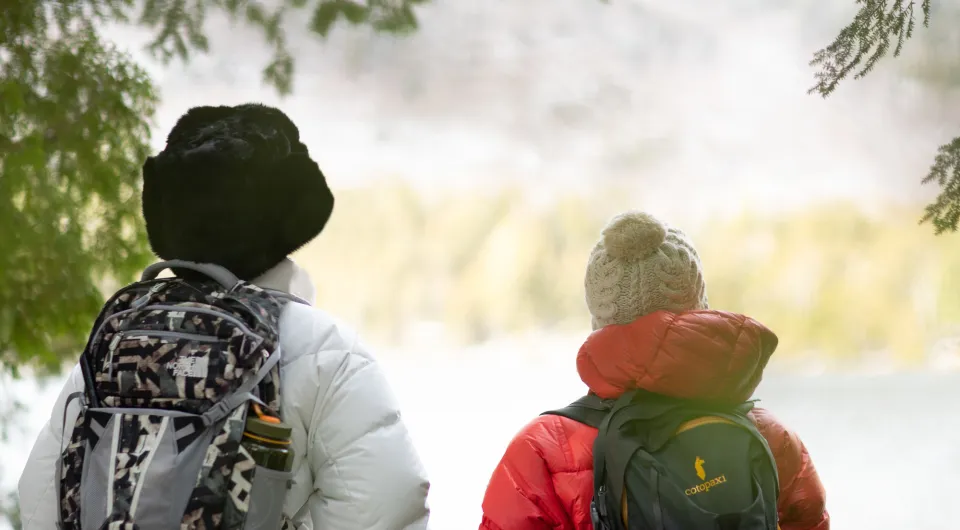 Two people in winter clothes with backpacks facing away looking over frozen pond