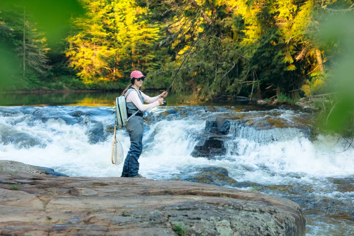 Woman stands on rocky shoreline in waders casting a fishing line in rushing water