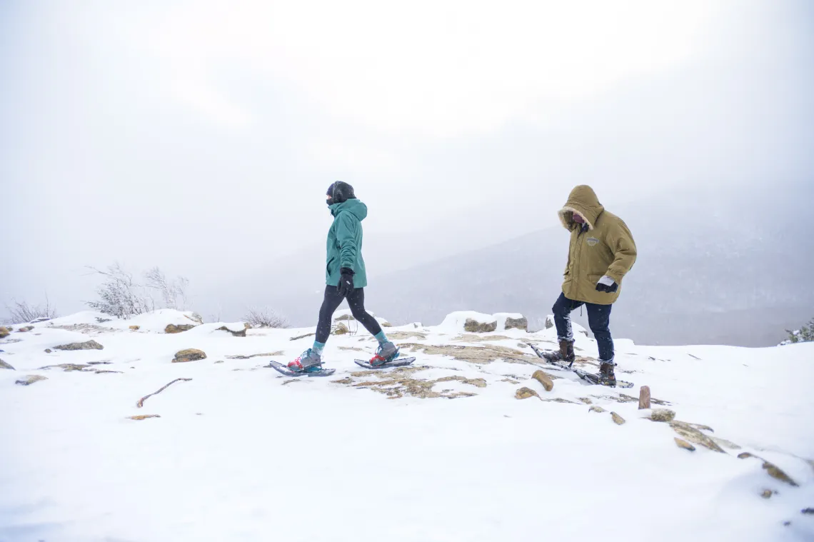 Two people in cold weather gear and snowshoes walk on snowy mountain summit