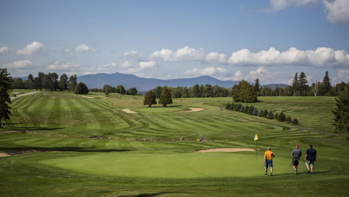 Three men appear small on a rolling, green golf course framed by trees and distant mountains.