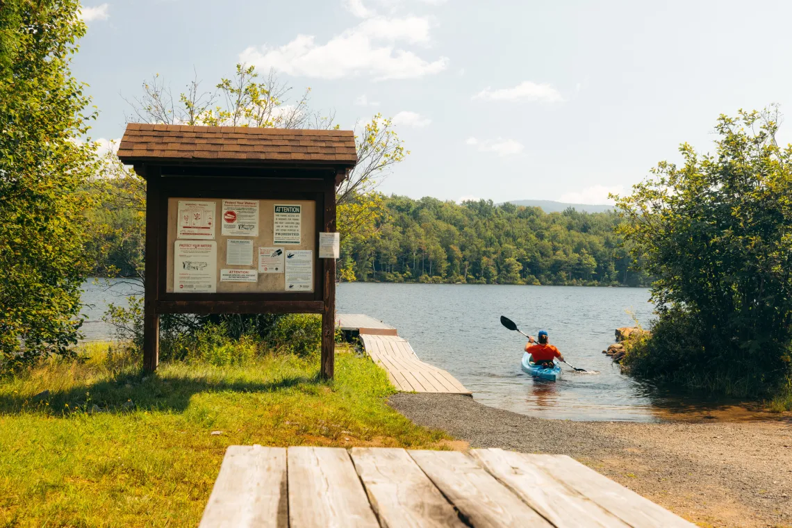 Man paddles away from shore in blue kayak next to a small dock and adjacent bulletin board