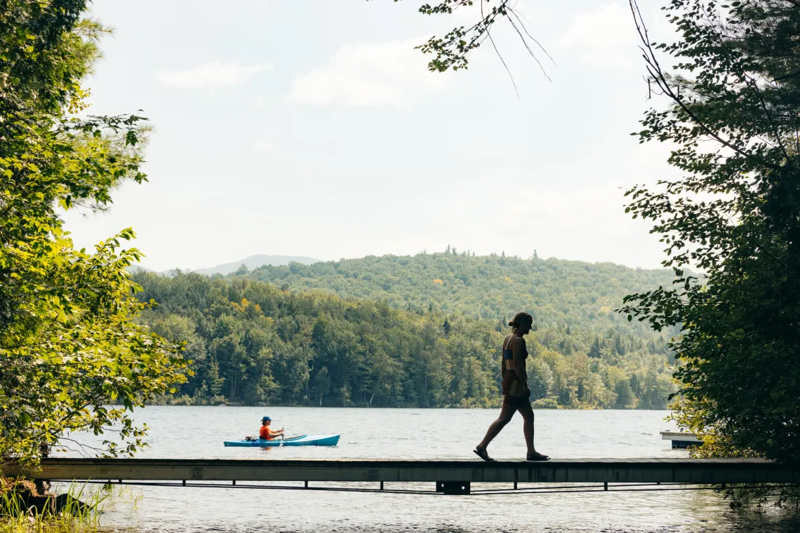 Silhouetted person walks along a boardwalk while a person sits in a blue kayak on the water behind them