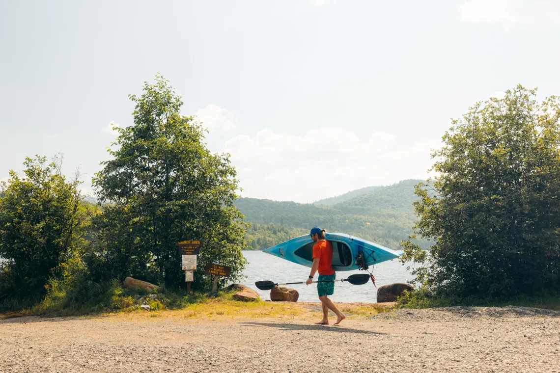 Man in t-shirt and shorts walks barefoot through lakeside parking area carrying a kayak on one shoulder and a paddle in the other hand